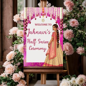 Könnte beinhalten: Ein rosa-weißes Willkommenszeichen mit floralen Verzierungen und einer Frau in traditioneller indischer Kleidung. Das Schild lautet "Willkommen zur Halb-Saree-Zeremonie von Jahnavi".