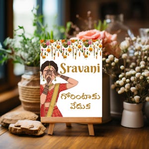 May include: A white sign on a wooden easel features the name "Sravani" in gold lettering, along with text in a different language. The sign has a floral border and an illustration of a woman in traditional Indian attire.