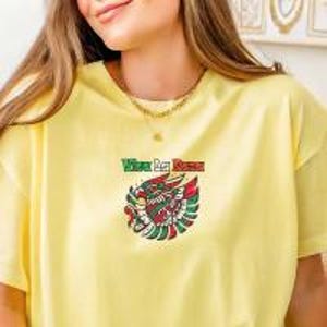 May include: A light yellow t-shirt with a colorful embroidered design of a bird with a banner that reads "Viva La Raza". The bird design features red, green, and white accents. A gold necklace is visible.
