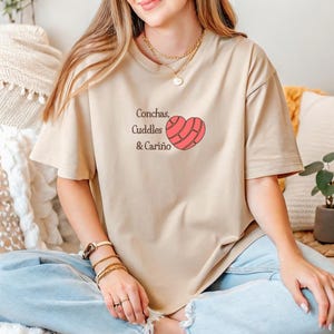 May include: Beige t-shirt with the text "Conchas, Cuddles & Cariño" and a heart-shaped concha design in red and pink. The shirt is worn with blue jeans. The overall theme is comfort and affection.
