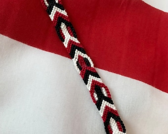 May include: A red, black, and white friendship bracelet with a chevron pattern.