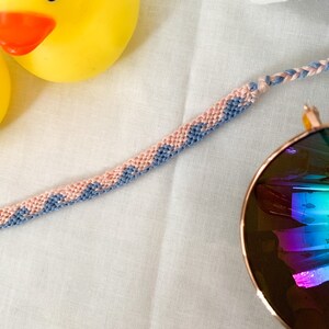May include: A pink and blue friendship bracelet with a knot at the end. The bracelet is lying on a white surface with a yellow rubber duck and sunglasses in the background.
