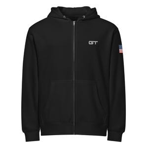 Op de afbeelding: Zwarte hoodie met ritssluiting, capuchon en voorzakken. De hoodie heeft een wit "GT"-logo op de borst en een kleine Amerikaanse vlag op de rechter mouw. De jas heeft een volledige ritssluiting en een capuchon met trekkoord.