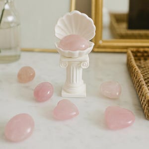 Può includere: Un supporto decorativo bianco a forma di conchiglia con una pietra rosa all'interno, sopra una colonna bianca. Diverse pietre di quarzo rosa di varie forme, tra cui ovali e cuori, sono disposte intorno.