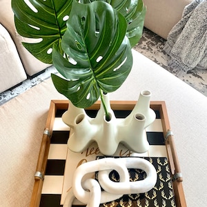 May include: A wooden tray with a black and white striped design. The tray features a white ceramic vase with a unique organic shape, a white chain link sculpture, and a black box with a gold deer antler pattern.