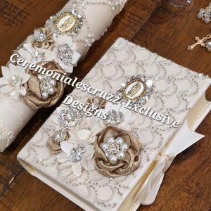 May include: A cream-colored candle and a matching book, both adorned with intricate silver beadwork, fabric flowers, pearl accents, and a central gold-toned oval medallion featuring a religious figure. The text reads Ceremonialescruzz Exclusive Designs.