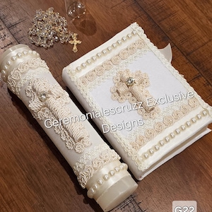 May include: Ivory-colored baptismal set featuring a decorated candle and a book, both adorned with crosses, pearls, and lace. The set includes a rosary and a small glass bottle. The text "Ceremonialescruzz Exclusive Designs" is visible.