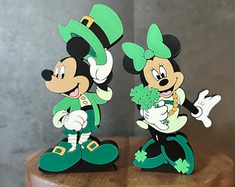 May include: Two wooden figurines of Mickey Mouse and Minnie Mouse dressed in green and white St. Patrick's Day outfits. Mickey is wearing a green hat and holding a shamrock. Minnie is wearing a green bow and holding a shamrock.