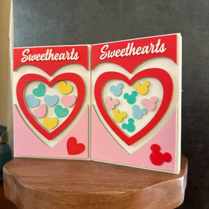 May include: Two pink and white cardboard boxes with the word "Sweethearts" printed on them. Each box has a red heart cutout with a different design inside. One heart has a pattern of colorful hearts, and the other has a pattern of colorful Mickey Mouse heads.