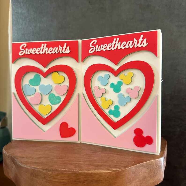 May include: Two pink and white cardboard boxes with the word "Sweethearts" printed on them. Each box has a red heart cutout with a different design inside. One heart has a pattern of colorful hearts, and the other has a pattern of colorful Mickey Mouse heads.