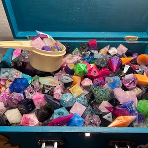 May include: A teal wooden box overflowing with a variety of colorful polyhedral dice. The dice are in various colors, including pink, blue, green, and orange. A small beige scoop is filled with dice.