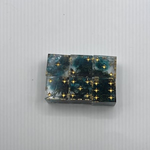 May include: Six square dice with a forest scene design. The dice have a teal and gold color scheme, with gold star-shaped markings. The dice are arranged in a rectangular shape.