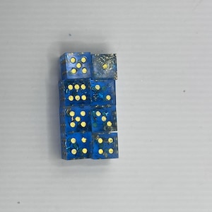 May include: A stack of seven blue dice with gold-colored pips. The dice are transparent with a glittery, textured appearance. The pips are arranged in the standard configuration for dice.