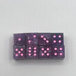 May include: A set of eight dice with a purple and silver glitter effect. The dice have pink dots indicating the numbers. The dice are arranged in two rows of four.
