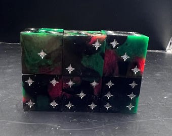 May include: Six square resin blocks, stacked in two rows, each with a unique blend of green, black, and red hues. Silver star-shaped accents are embedded on each face. The blocks are arranged on a dark surface.