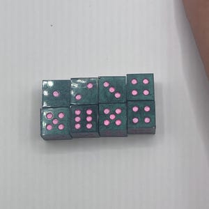 May include: A collection of eight green dice with pink dots arranged in two rows. Each die displays a different number of dots, ranging from one to six. The dice are set against a plain white background.