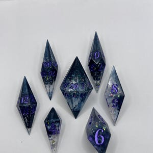 Op de afbeelding: Een set van zeven veelzijdige dobbelstenen met een scherpe, langwerpige ruitvorm. De dobbelstenen zijn gemaakt van doorschijnende donkerblauwe en witte hars met ingebedde glitters en hebben dieppaarse cijfers in het oppervlak gegraveerd.