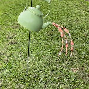 May include: A light green metal teapot garden stake with a handle and spout. The spout appears to be pouring out a stream of pink, orange, and clear beads. The teapot is mounted on a black metal pole.