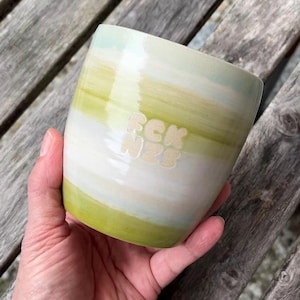 Könnte beinhalten: Ein Keramikbecher mit horizontalen Streifen in hellgrüner, blassgelber und weißer Glasur. Die Oberfläche hat ein glattes, glänzendes Finish mit dem Text FCK NZS, der in einer blockartigen serifenlosen Schrift in der Mitte eingeprägt ist.