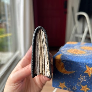 May include: A black leather-bound journal with a visible stack of pages. The journal is held in a hand, showing the spine and the pages.