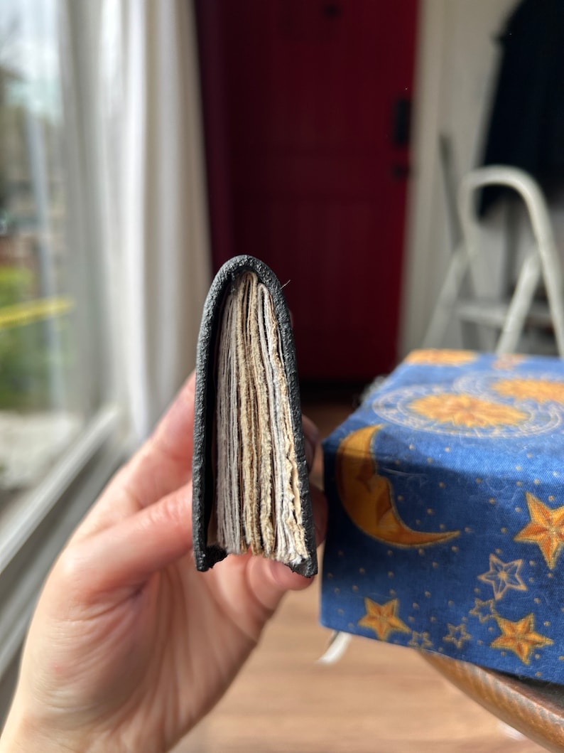 May include: A black leather-bound journal with a visible stack of pages. The journal is held in a hand, showing the spine and the pages.