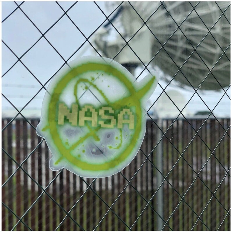 May include: A clear sticker featuring the NASA logo. The logo is a pixelated design with the word "NASA" in gold letters. The logo is surrounded by a green and yellow splatter design. The sticker is adhered to a window with a wire fence in front.