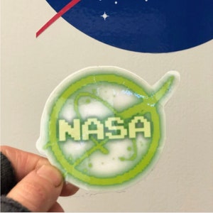 May include: A clear sticker featuring the NASA logo in green and white. The sticker has a pixelated design and the word "NASA" is prominently displayed. The background shows a portion of the NASA logo.