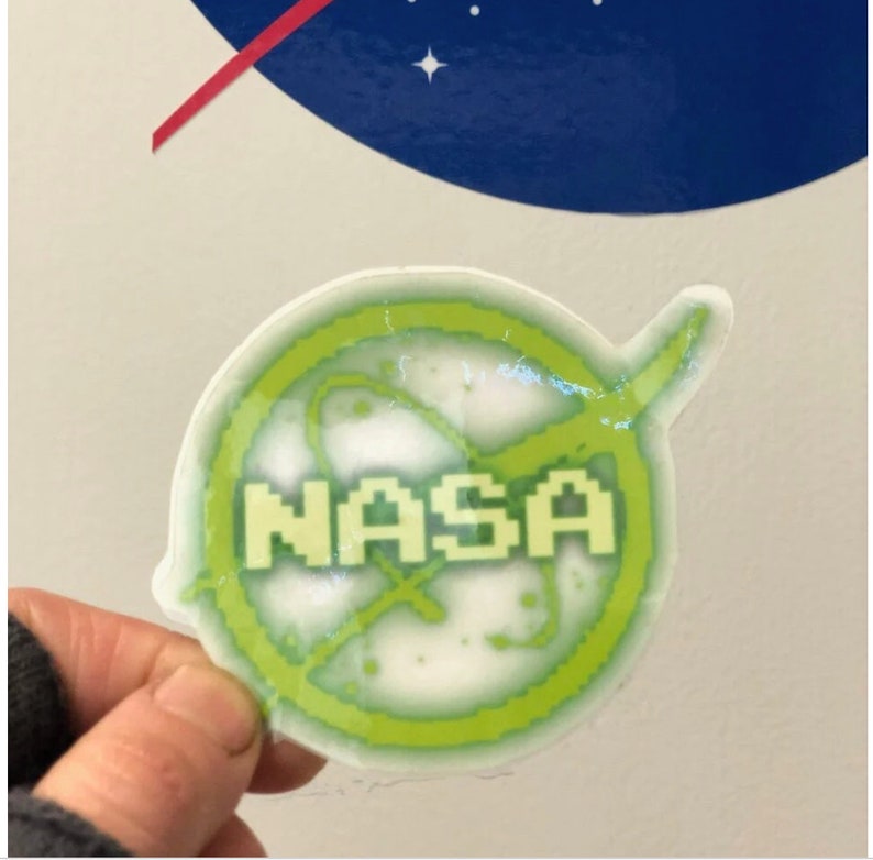 May include: A clear sticker featuring the NASA logo in green and white. The sticker has a pixelated design and the word "NASA" is prominently displayed. The background shows a portion of the NASA logo.