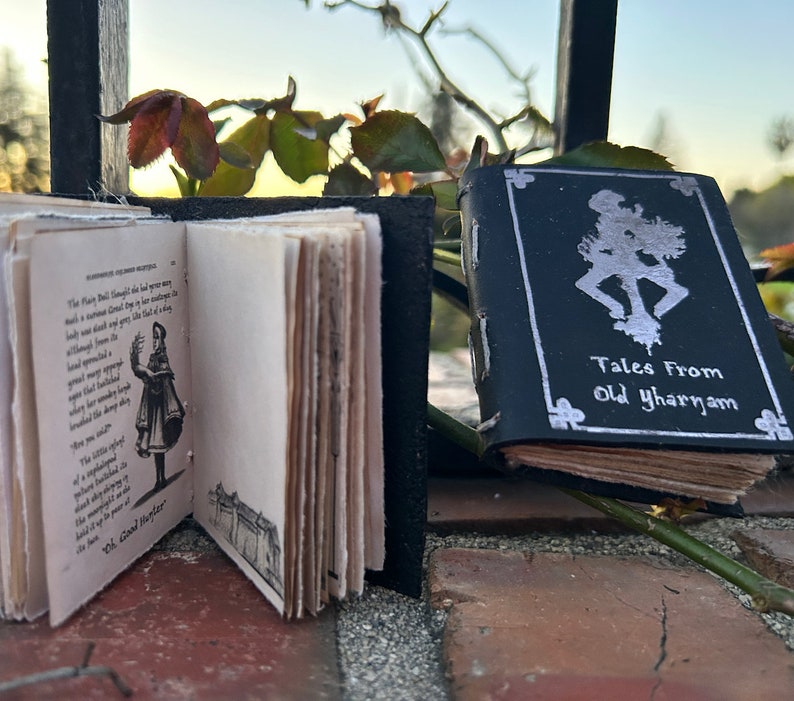 May include: Two small, leather-bound books with silver lettering. The book on the left is open to a page with a black and white illustration of a girl in a dress. The book on the right has the title "Tales From Old Yharnam" on the cover.
