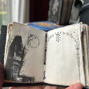 May include: A small black leather-bound journal with a hand-drawn illustration of a house and a moon on the front cover. The back cover has a decorative design with a symbol and floral elements.