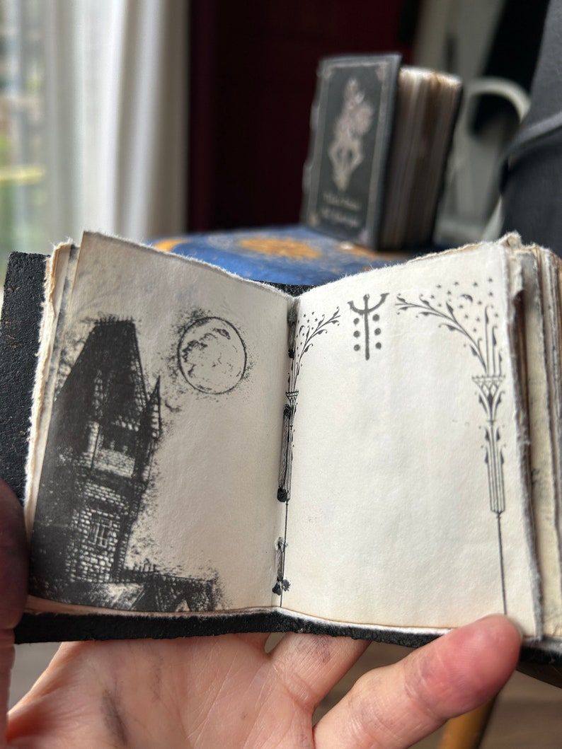May include: A small black leather-bound journal with a hand-drawn illustration of a house and a moon on the front cover. The back cover has a decorative design with a symbol and floral elements.