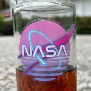 May include: A clear glass jar with a NASA logo sticker. The sticker features a pink and purple background with a blue neon-style arc and the NASA text in white. The jar contains a dark amber liquid.
