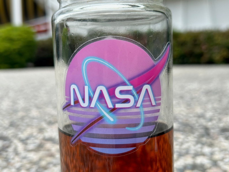 May include: A clear glass jar with a NASA logo sticker. The sticker features a pink and purple background with a blue neon-style arc and the NASA text in white. The jar contains a dark amber liquid.