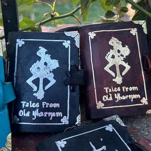 May include: Two small black leather-bound journals with silver and gold accents. The covers feature a stylized symbol and the text "Tales From Old Yharnam".