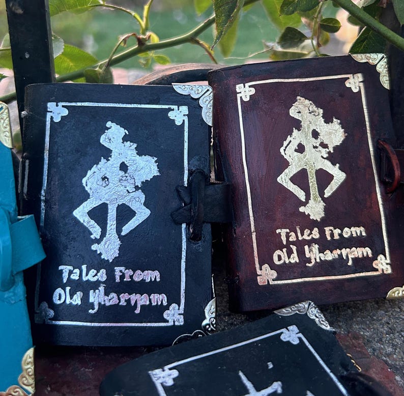 May include: Two small black leather-bound journals with silver and gold accents. The covers feature a stylized symbol and the text "Tales From Old Yharnam".