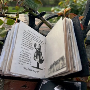 May include: A small, black leather-bound book with a white page open to a black and white illustration of a woman in a long dress holding a flower. The text on the page reads "The Main Dall thought he had never seen such a curious Great sight in his entire existence. Lady, some sleek and grey, like that of a dove, although from its head sprouted many appendages. When her wooden ladle brushed the damp shin..." and "Oh, Good Hunter".