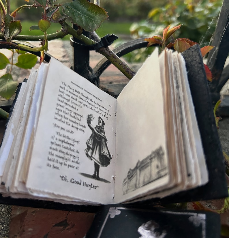 May include: A small, black leather-bound book with a white page open to a black and white illustration of a woman in a long dress holding a flower. The text on the page reads "The Main Dall thought he had never seen such a curious Great sight in his entire existence. Lady, some sleek and grey, like that of a dove, although from its head sprouted many appendages. When her wooden ladle brushed the damp shin..." and "Oh, Good Hunter".