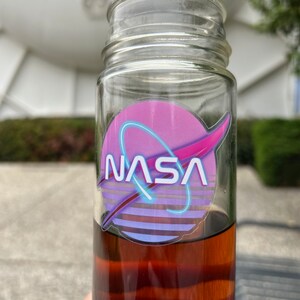 May include: A clear glass bottle filled with a dark amber liquid. The bottle features a NASA logo sticker with a pink and purple design and the word "NASA" in white. The bottle is outdoors with a blurred background.