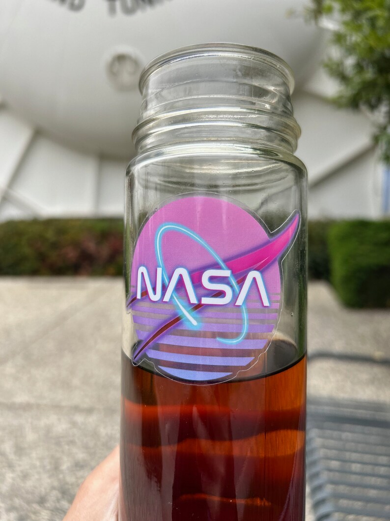 May include: A clear glass bottle filled with a dark amber liquid. The bottle features a NASA logo sticker with a pink and purple design and the word "NASA" in white. The bottle is outdoors with a blurred background.