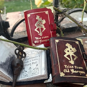 May include: Two small, leather-bound books with gold lettering and illustrations. The books are titled "Tales From Old Yharnam". One book is red and the other is brown. A small, antique key is lying on top of a white book.