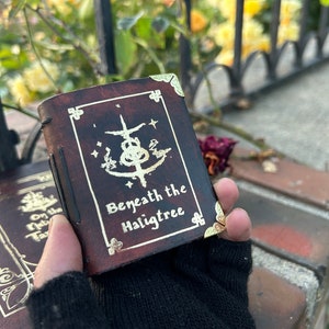 May include: A brown leather-bound journal with gold accents and the text "Beneath the Hailgtree" on the front cover. The journal has a decorative design with a symbol and stars.