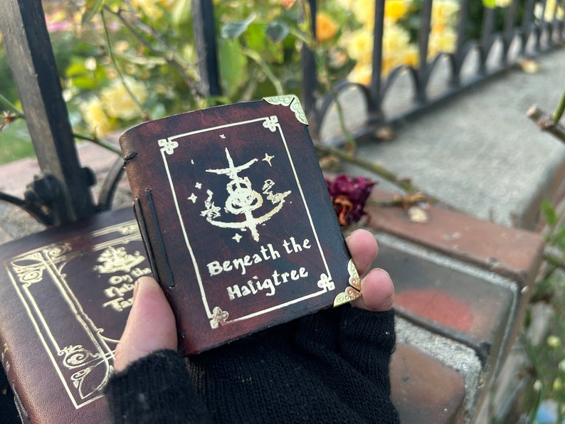May include: A brown leather-bound journal with gold accents and the text "Beneath the Hailgtree" on the front cover. The journal has a decorative design with a symbol and stars.