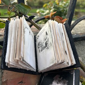 May include: A small black leather-bound journal with a hand-drawn illustration of a house and trees on the open page. The journal is closed and has a worn, vintage look.
