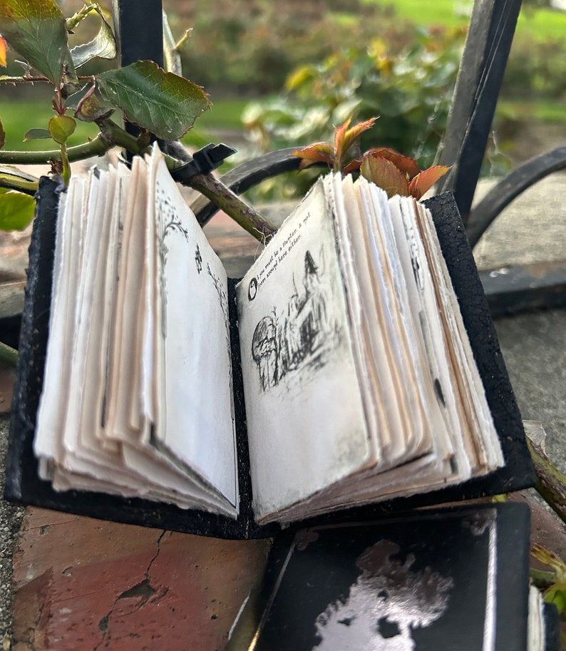 May include: A small black leather-bound journal with a hand-drawn illustration of a house and trees on the open page. The journal is closed and has a worn, vintage look.