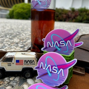 May include: A clear glass bottle with a NASA logo sticker and a toy truck with a USA flag. The NASA logo is a pink and purple circle with a blue neon outline. The toy truck has the text "USA 1096" and "NASA United States".