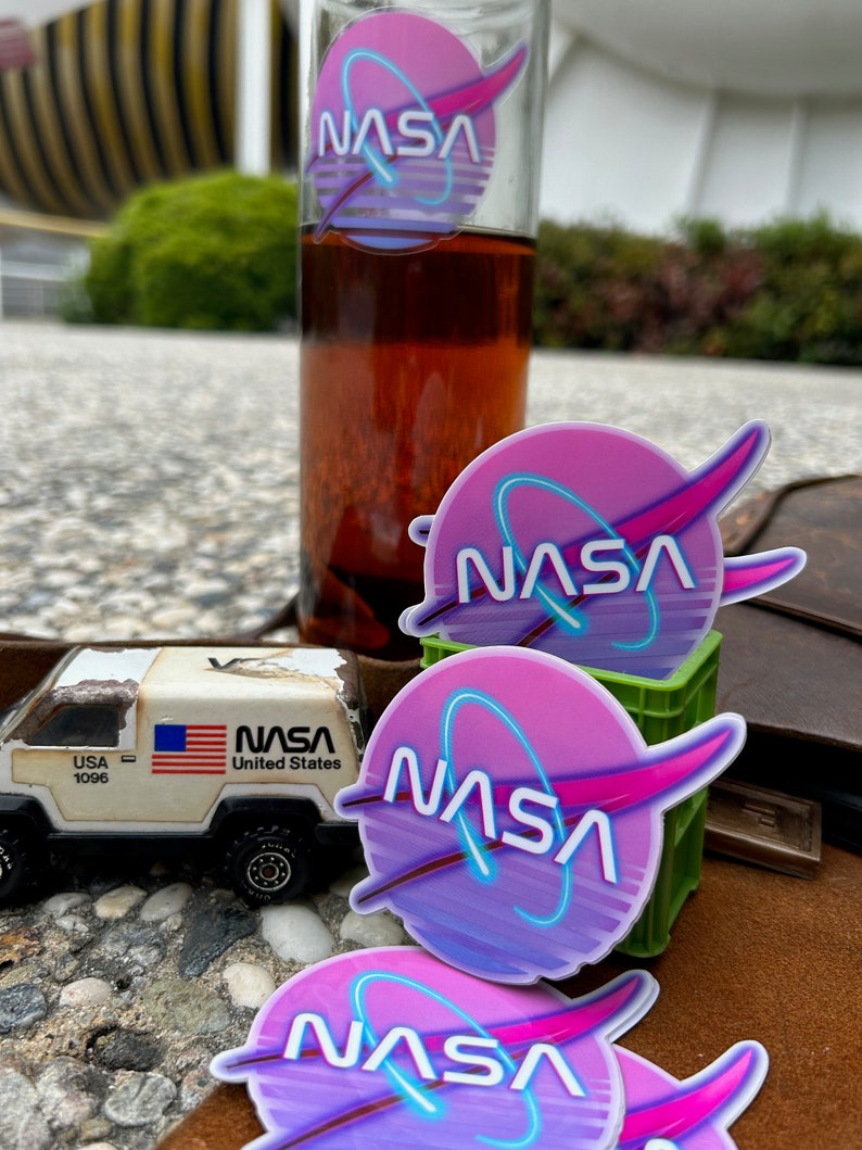 May include: A clear glass bottle with a NASA logo sticker and a toy truck with a USA flag. The NASA logo is a pink and purple circle with a blue neon outline. The toy truck has the text "USA 1096" and "NASA United States".