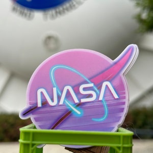 May include: A sticker featuring the NASA logo in a retro style. The design includes a pink and purple color scheme with a blue neon-style outline. The word "NASA" is prominently displayed in white. The sticker is on a green crate.