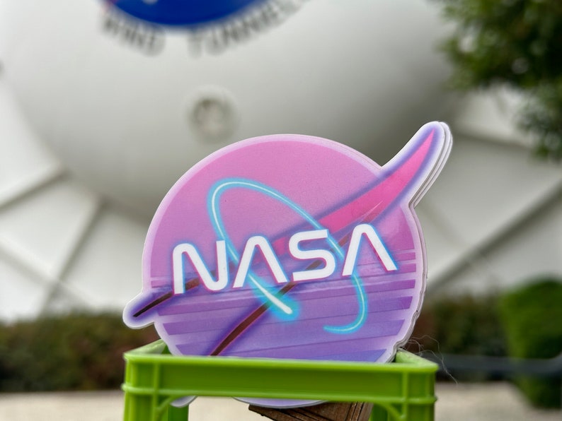 May include: A sticker featuring the NASA logo in a retro style. The design includes a pink and purple color scheme with a blue neon-style outline. The word "NASA" is prominently displayed in white. The sticker is on a green crate.