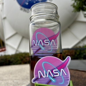 May include: A clear glass bottle with a brown liquid inside and a NASA logo sticker. A second NASA logo sticker sits in a small green crate. The background features a large white structure with the NASA logo.