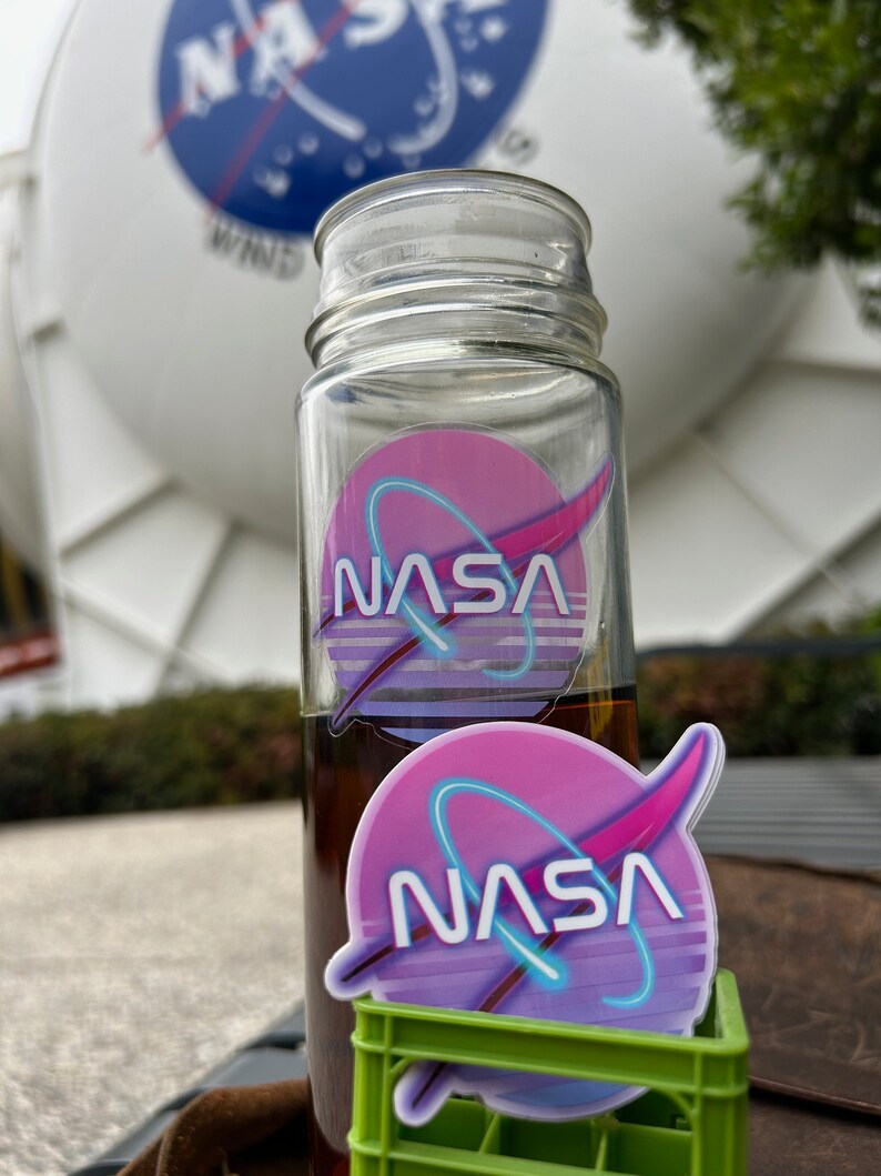 May include: A clear glass bottle with a brown liquid inside and a NASA logo sticker. A second NASA logo sticker sits in a small green crate. The background features a large white structure with the NASA logo.